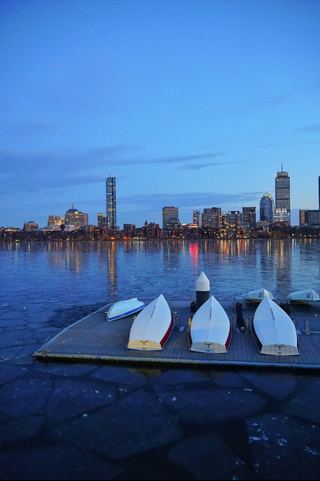 Sailboats hauled onto a dock beside the frozen Charles River with the Back Bay skyline at dusk