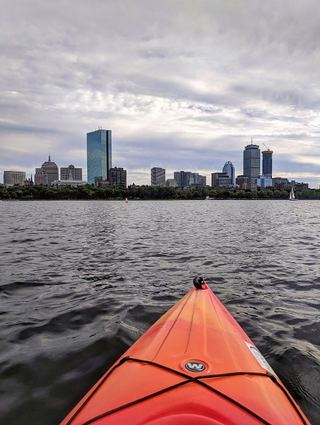 The bow of a kayak on the Charles River with the Boston skyline beyond